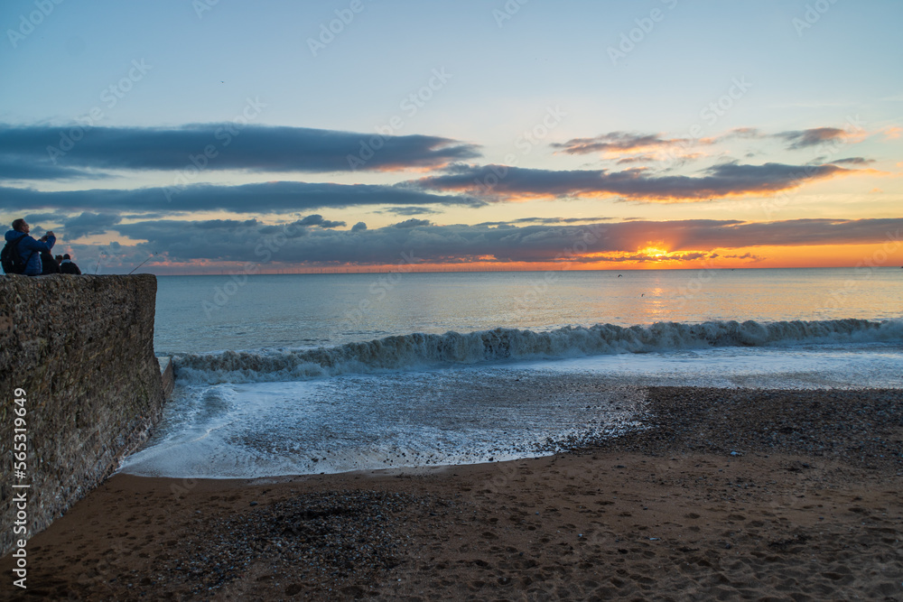 People watch waves and surf on the beach with clouds and setting sun over the English Channel in the background. Brighton, UK