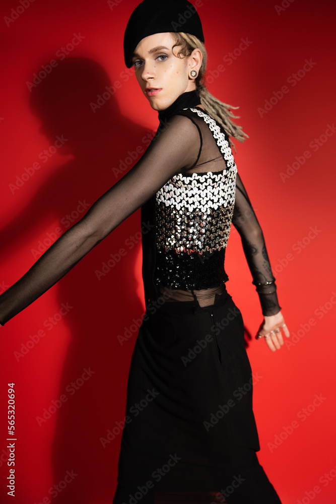 Fototapeta premium young nonbinary model in elegant top with sequins looking at camera on red with shadow.