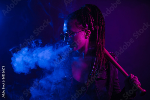A young woman with dreadlocks smokes a cigarette in a club. Wearing black glasses, he holds a painless bat in his hands. Girl with afro pigtails in the smoke at night in neon color. Scoundrel