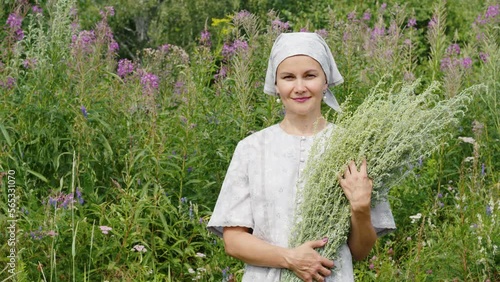 Village woman holds armful of wormwood in hands, looking into camera. Portrait of cute farm girl in headscarf with wermout among tall grasses. Attractive lady keeps absinthium herbs near wildflowers photo