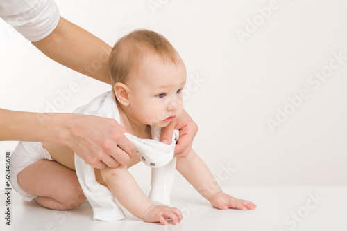 Young adult mother hands putting white bodysuit on crawling baby boy on floor at light gray wall background. Closeup. Side view. Parent daily duties. 8 old months infant.