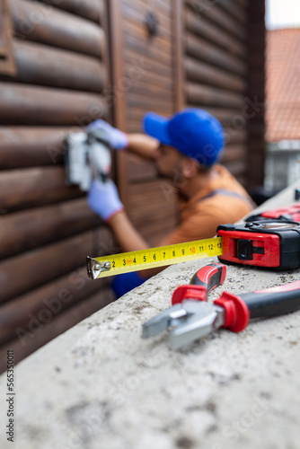 Close up of measuring tape and pliers on concrete fence with professional worker renovating wooden house in the background.