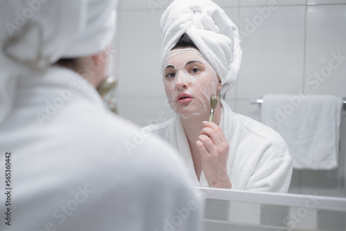 Beautiful young woman doing facial massage with gua sha tool in front of mirror at home