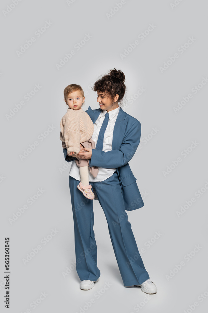 full length of happy businesswoman in blue suit standing and holding toddler daughter on grey. full length of happy businesswoman in blue suit standing and holding toddler daughter on grey.