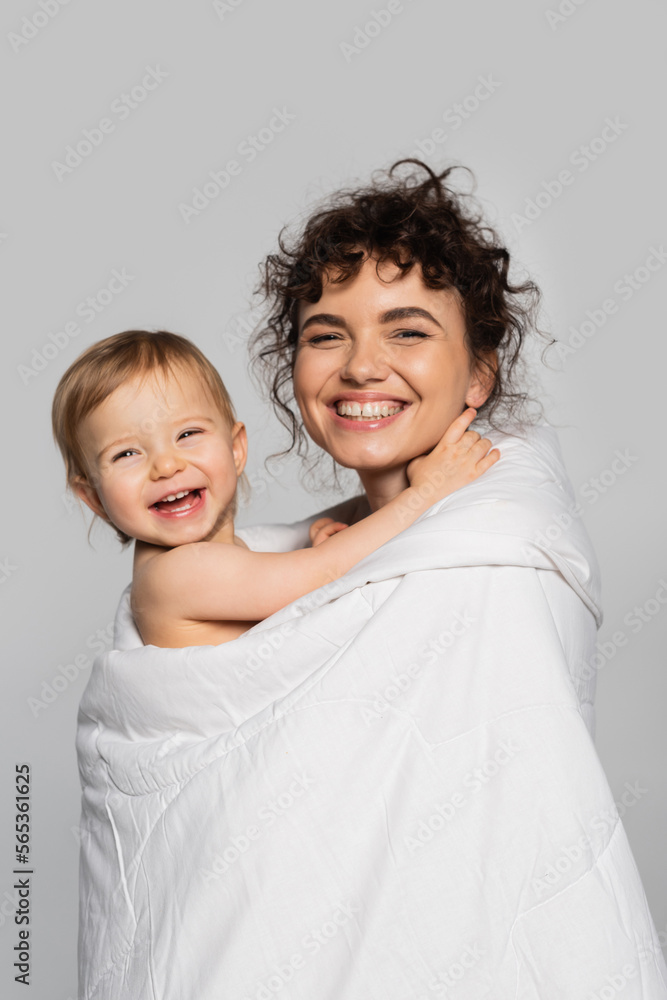 happy mother and cheerful toddler daughter covered in duvet isolated on grey.