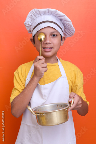 Happy smiling little boy in apron with saucepan and spoon over orange background