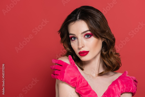 portrait of young woman with magenta color eye shadow standing with crossed arms while embracing herself isolated on pink.