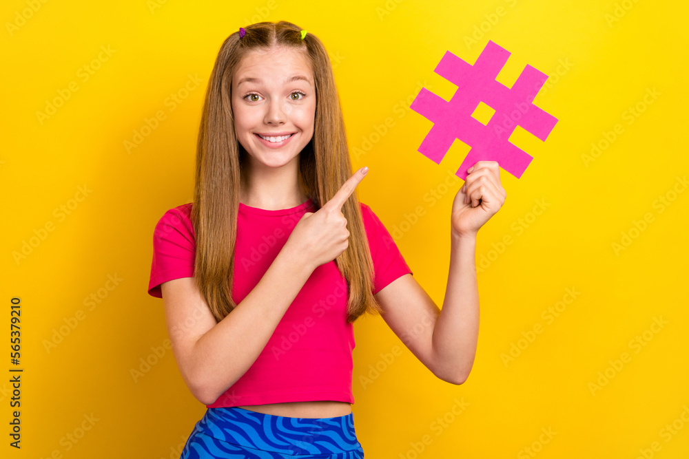 Photo of young teenager schoolgirl wear stylish pink crop top finger directing paper hashtag symbol popular pin isolated on yellow color background