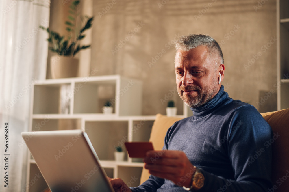 Middle aged man using a laptop and a credit card while sitting on a sofa at home Middle aged man using a laptop and a credit card while sitting on a sofa at home