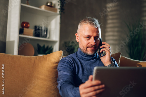 Middle aged man using a smartphone and a laptop while sitting on a sofa at home