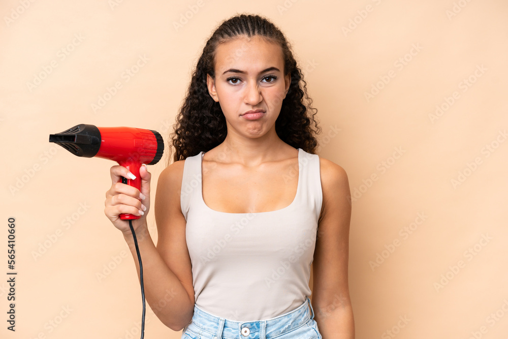 Young woman holding a hairdryer isolated on beige background with sad expression Young woman holding a hairdryer isolated on beige background with sad expression