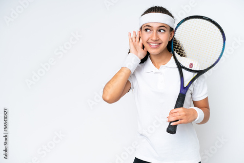 Young tennis player woman isolated on white background listening to something by putting hand on the ear © luismolinero