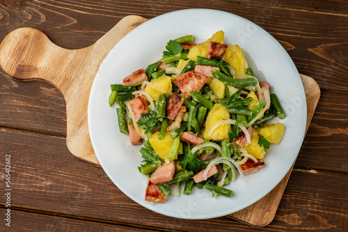 Liege potato salad with beans and ham and bacon, traditional Belgian salad in a white plate on a wooden board, top view.