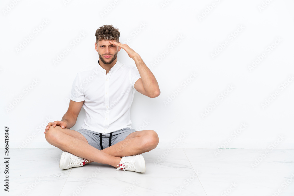 Young blonde man sitting on the floor isolated on white background looking far away with hand to look something