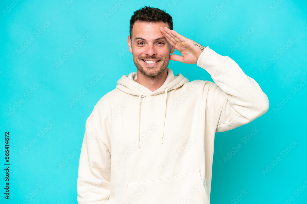 Young caucasian handsome man isolated on blue background saluting with hand with happy expression