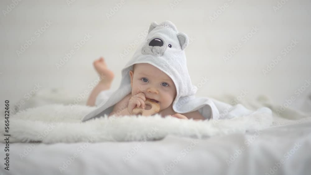 A cute funny little baby with a wood teether is resting in bed after bathing, covered with a towel, laughing happily. The concept of a baby bath. Copy space. Soft focus. A cute funny little baby with a wood teether is resting in bed after bathing, covered with a towel, laughing happily. The concept of a baby bath. Copy space. Soft focus.