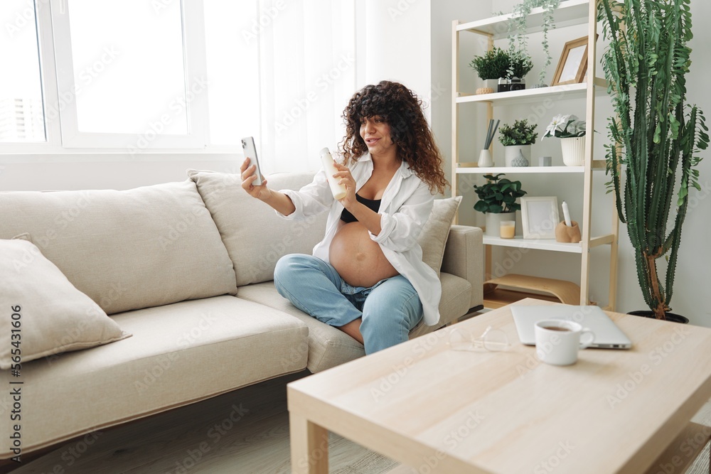 Pregnant woman smile blogger showing baby feeding bottle filming herself on phone, video chat sitting on sofa at home freelancer in last month of pregnancy Pregnant woman smile blogger showing baby feeding bottle filming herself on phone, video chat sitting on sofa at home freelancer in last month of pregnancy