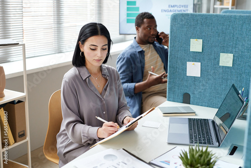 Tableau sur toile Portrait of young businesswoman writing on clipboard while working in office cub