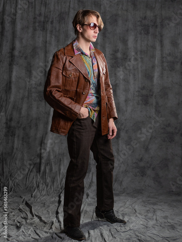 confident dude in a colorful shirt and brown leather jacket, a portrait in sunglasses in the studio on a gray background
