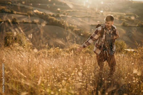 Happy man walking and carrying his backpack and hiking. He is smiling and touching the grass. He is going through a grass meadow.