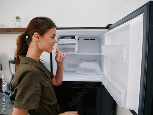 Woman smiling with teeth looking into camera in kitchen at home opened freezer empty with ice inside  home refrigerator  defrosted  view from back  stylish interior.