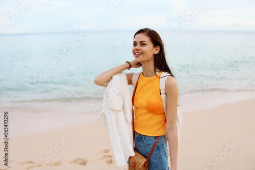 Woman smile with teeth after swimming in the ocean with a backpack in a wet yellow tank top and denim shorts walks along the beach, summer vacation on an island by the ocean in Bali sunset