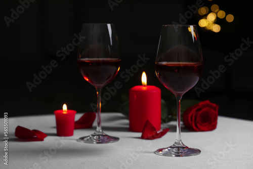 Glasses of red wine, rose flower and burning candles on white table against blurred lights. Romantic atmosphere