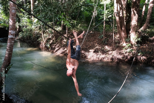 A tourist (middle aged man) has fun hanging upside down on a rope above crystal clear creek in the jungle on sunny day. Krabi Province, Thailand.