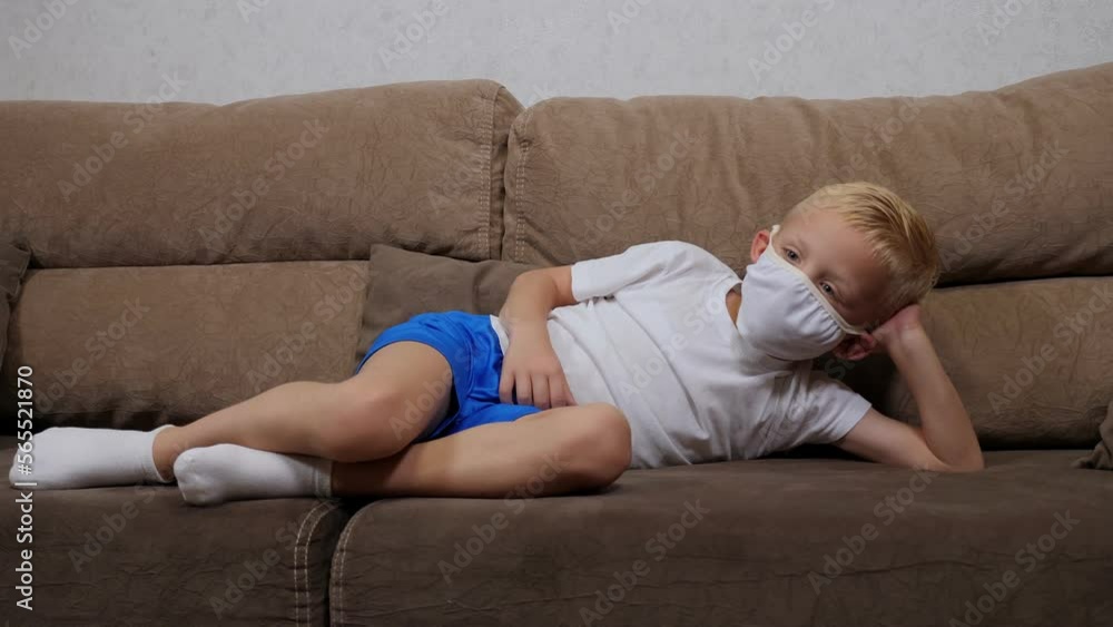 A small boy in a medical mask is lying on the sofa at home, holding a remote control in his hands and switching channels. Children on distance learning during quarantine. A small boy in a medical mask is lying on the sofa at home, holding a remote control in his hands and switching channels. Children on distance learning during quarantine.