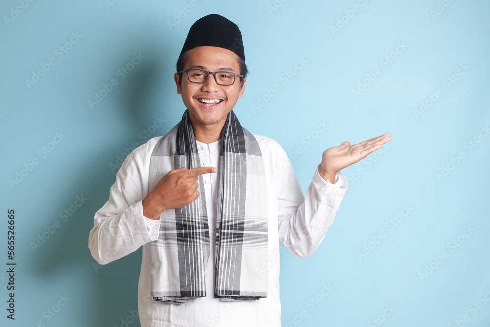 Portrait of young Asian muslim man showing product and pointing with his hand and finger to the side. Isolated image on blue background Portrait of young Asian muslim man showing product and pointing with his hand and finger to the side. Isolated image on blue background