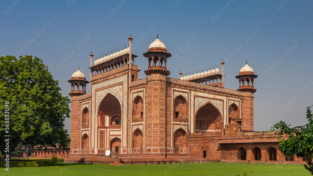 The ancient mosque Kau Ban in the Taj Mahal complex is built of red sandstone and white marble. Arches, towers, domes, inlays of precious stones are visible.  Green vegetation around. Blue sky. India