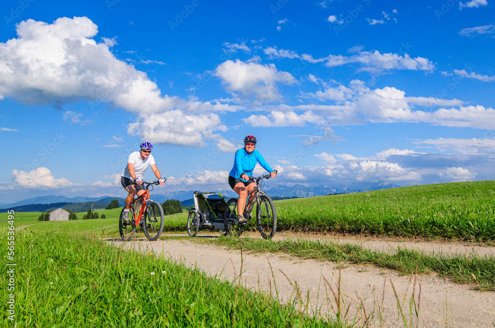 Obraz premium Eltern mit Nachwuchs im Buggy unterwegs mit dem Mountainbike, die Voralpen-Landschaft im Allgäu genießen 