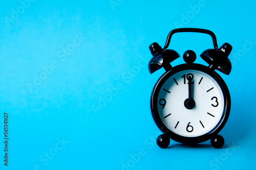 Black alarm clock isolated on blue background. The clock set at 12pm. Copy space and time concept photo