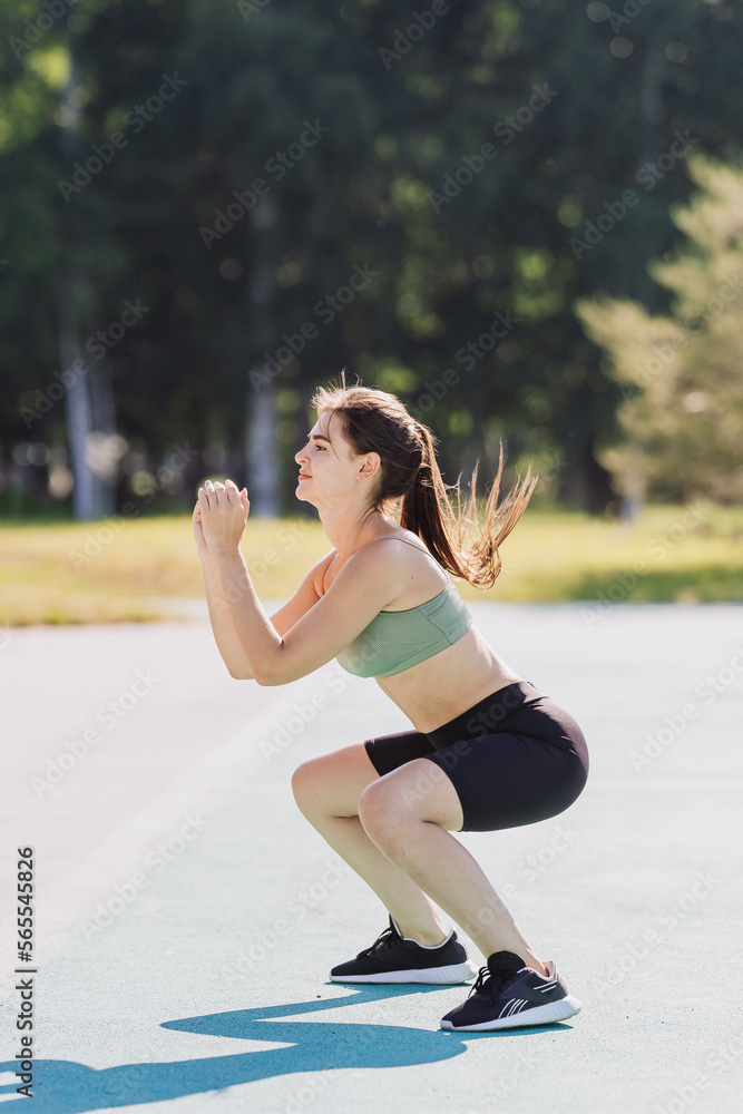 Vertical shoot of young student girl in sportswear making squats outdoors. Brunette American fit student training at park. Workout, fitness people, youth. Healthy lifestyle. Sporty girl at exercise. Vertical shoot of young student girl in sportswear making squats outdoors. Brunette American fit student training at park. Workout, fitness people, youth. Healthy lifestyle. Sporty girl at exercise.