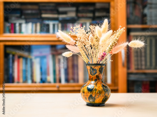 side view of bouquet of dried plants in vase on light brown wooden table with bookcase on background photo