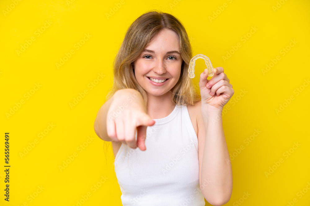 Blonde English young girl holding invisible braces isolated on yellow background points finger at you with a confident expression Blonde English young girl holding invisible braces isolated on yellow background points finger at you with a confident expression