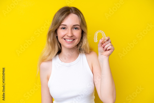 Blonde English young girl holding invisible braces isolated on yellow background smiling a lot