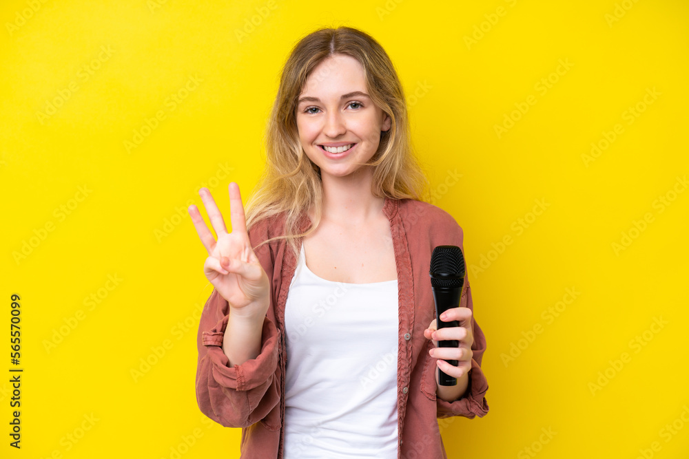 Young singer caucasian woman picking up a microphone isolated on yellow background happy and counting three with fingers Young singer caucasian woman picking up a microphone isolated on yellow background happy and counting three with fingers