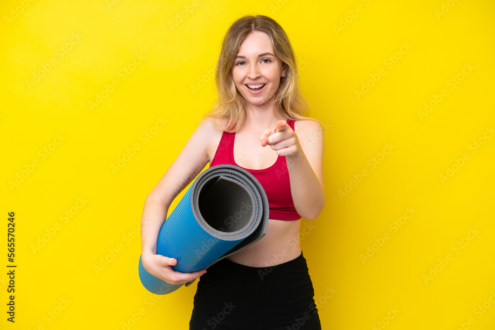 Young sport caucasian woman going to yoga classes while holding a mat isolated on yellow background surprised and pointing front Young sport caucasian woman going to yoga classes while holding a mat isolated on yellow background surprised and pointing front