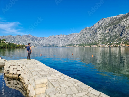 Hiking man in small port in Kotor Bay in coastal town Prcanj, Adriatic Mediterranean Sea, Montenegro, Balkans, Europe. Scenic view on village Dobrota. Fjord winding along beautiful Dinaric Alps