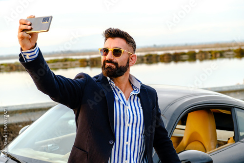 Handsome, elegant, bearded man takes a picture of himself right next to his sports car.
