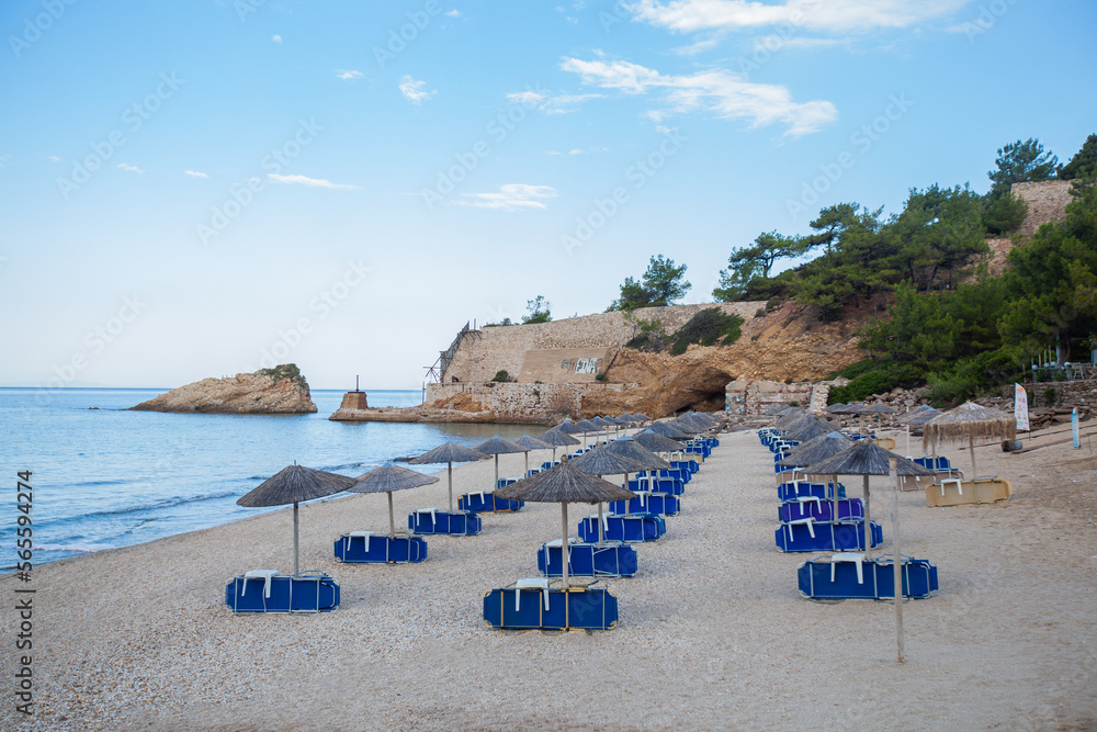Empty sand beach with sunbeds and parasols, surround rocks, in calm summers morning. Blue sky with clouds. Summer vacation.	
