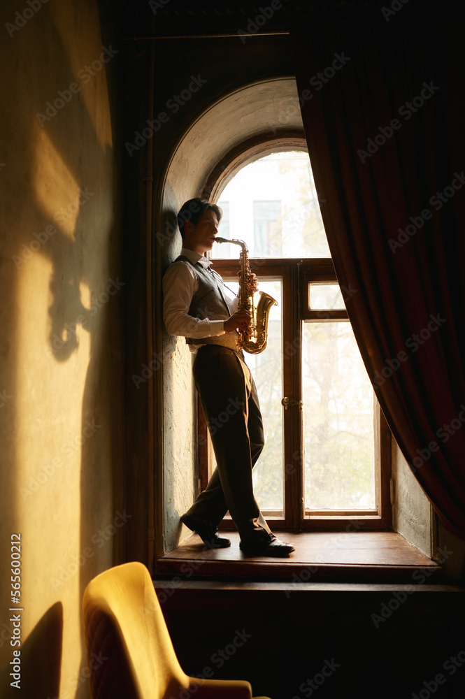 Creative young man playing saxophone standing on windowsill Creative young man playing saxophone standing on windowsill