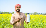 Confident young farmer showing fertilizer bottle by pointing finger while looking at camera at farmland - concept of product recommendation, agricultural and promotion.