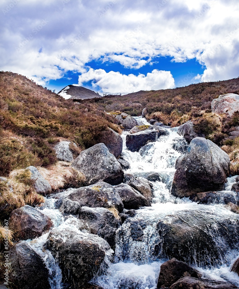 Obraz premium Stream rushing over rocks in Cairngorms National Park, Scotland, popular hiking and skiing area. A tall peak rises in the distance, snow on one face.
