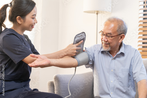 Pressure measurement of hypertension in elderly man sitting on couch, young woman nurse or caregiver care checking, measuring heart, blood by using sphygmomanometer monitor at home after retirement. photo