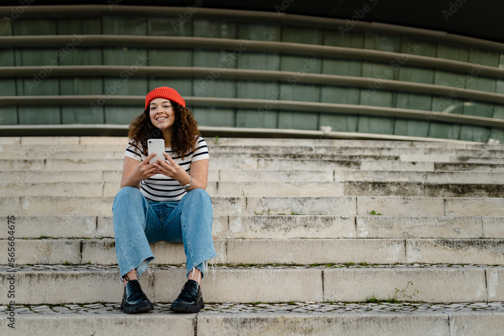 pretty curly smiling woman sitting in city street in striped t-shirt and knitted red hat, using smartphone