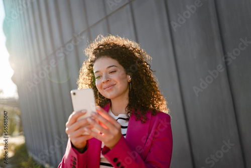 pretty curly smiling woman walking in city street in stylish jacket  using smartphone