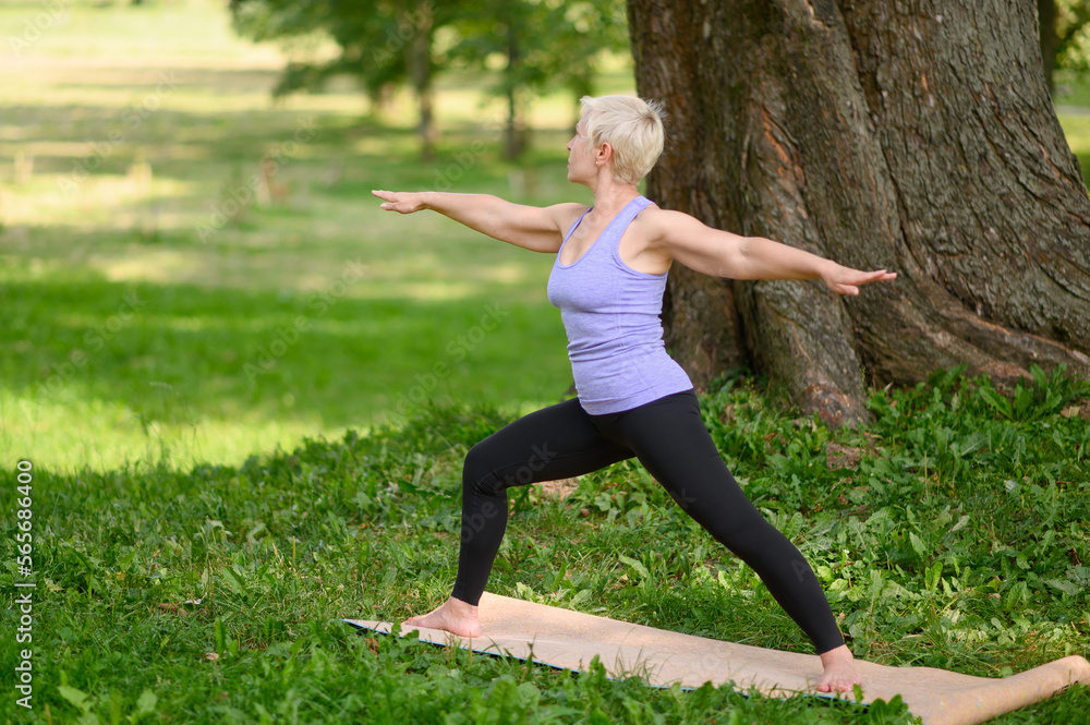 Fototapeta premium Middle aged caucasian woman in sportswear doing yoga outdoors in warrior pose, Virabhadrasana II. The concept of yoga, stretching, healthy lifestyle. Sports activities in nature. Copy space.