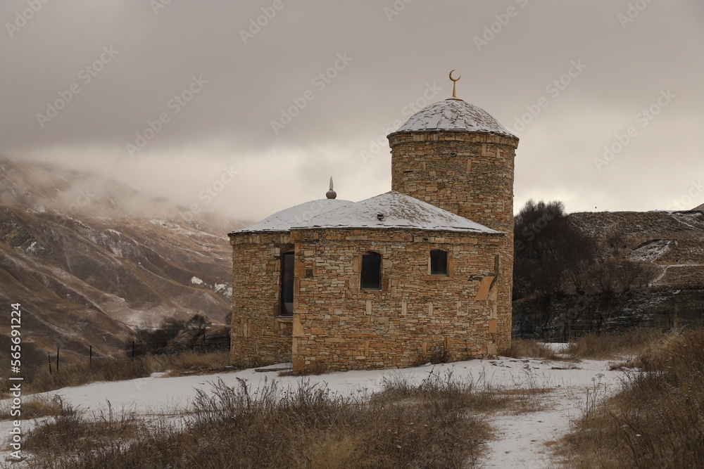 An ancient stone Muslim temple in the mountains of Dagestan - stands on a snowy slope against the backdrop of mountains and a gloomy sky.